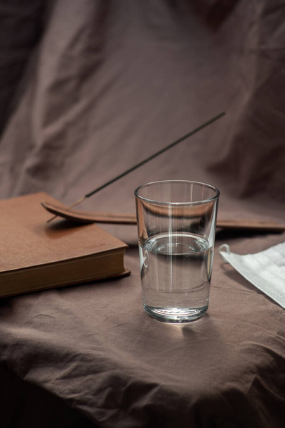 Композиція являє собою скляну чашку наполовину наповнену водою, книгою, маскою та димом на тлі тканини. М'яке світло. Колір трендів
 - Фото, зображення
