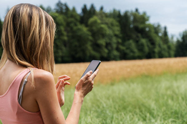 Młoda kobieta stojąca w lecie charakter pól i łąk przeglądających się na jej telefon komórkowy. - Zdjęcie, obraz
