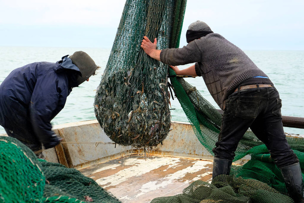 Fischer bei der Arbeit. Matrosen ziehen das Schleppnetz mit dem Fang auf das Deck des Schiffes. Fisch im Netz gefangen. Angeln. Schwarzes Meer. Überwiegend bewölkt. - Foto, Bild