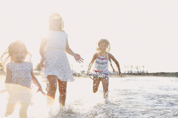 Little girls in colorful dresses having fun and running in sea water while enjoying summer holidays on beach in sunny day. Holiday and happiness concept - Photo, Image