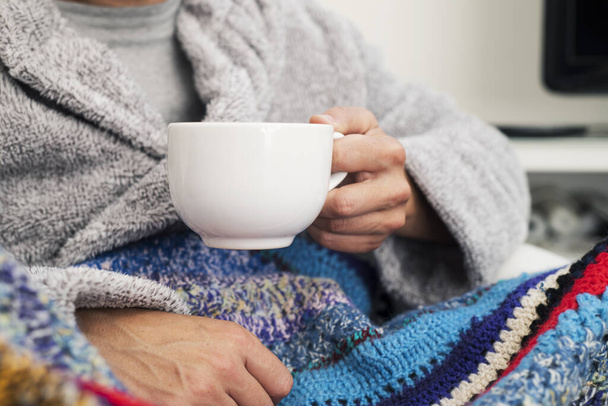 Крупный план больного кавказца дома, одетого в пушистый серый халат и завернутого в красочное трикотажное одеяло, согретого чашкой горячего супа
 - Фото, изображение