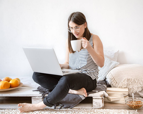 Μια νεαρή γυναίκα δουλεύει εξ αποστάσεως σε έναν υπολογιστή στο σπίτι. Ελεύθερος επαγγελματίας και εργασία στο Διαδίκτυο. - Φωτογραφία, εικόνα