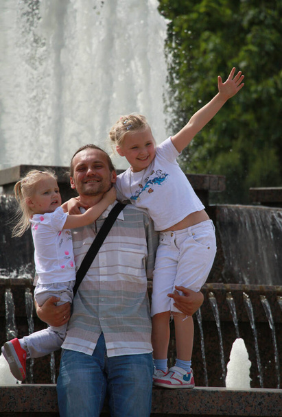 Ο πατέρας κρατά δύο κόρες στην αγκαλιά του στο βάθος του σιντριβανιού. - Φωτογραφία, εικόνα
