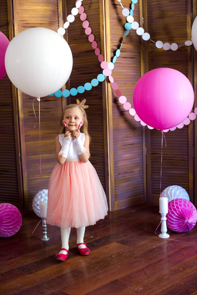 Μικρό χαριτωμένο κορίτσι με ξανθά μαλλιά σε ένα ροζ φόρεμα και μια πριγκίπισσα στέμμα με μεγάλα φωτεινά μπαλόνια και γιρλάντες. Παιδικές διακοπές. Παιδική διακόσμηση. Μπαλόνια - Φωτογραφία, εικόνα