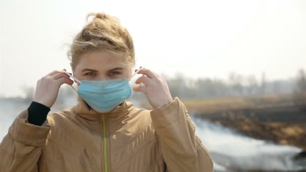 Zdenerwowana dziewczyna zakłada maskę medyczną. Pole do palenia. Spalona trawa. Zanieczyszczenie środowiska. Zwolnij trochę. Zbliżenie - Materiał filmowy, wideo
