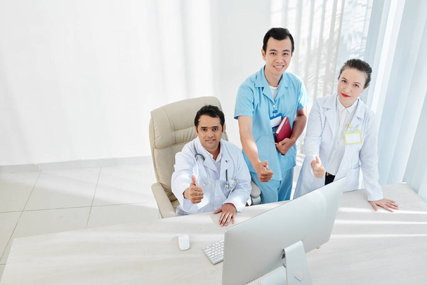 Monietninen ryhmä lääkäreitä toimistotiskillä näyttää peukalot ylöspäin ja hymyilee kameralle
 - Valokuva, kuva