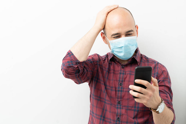 彼のスマートフォンを見て外科用マスクを持つ白人のはげ男の肖像とcovid19コロナウイルスについての悪いニュースを読む - 写真・画像