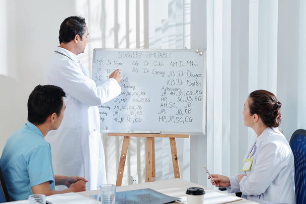 Médecin en chef discutant du calendrier de chirurgie sur tableau blanc avec ses collègues lors de la réunion au bureau
 - Photo, image