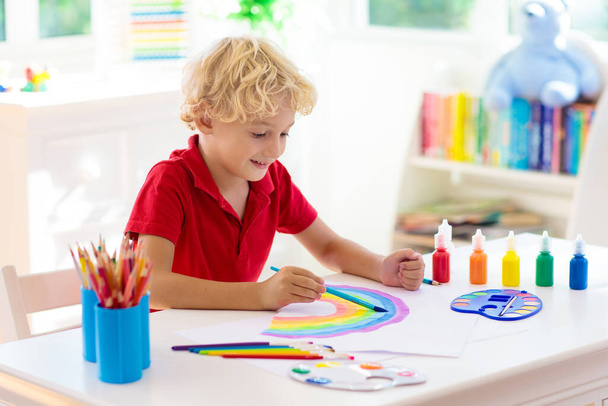 Kinderen schilderen. Quarantaine plezier. Een kind dat regenboog schildert, teken van hoop tijdens een coronavirus uitbraak. Kleine jongen tekening. Een schooljongen die huiswerk maakt. Kunstnijverheid voor kinderen. Verf op handen van kinderen.  - Foto, afbeelding
