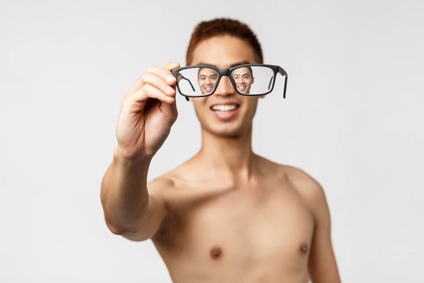 Krása, lidé a životní styl. Detail portrét nadšeného asijského muže s nahým trupem, ukazující brýle, dívající se přes brýle a usmívající se pozitivní, stojící bílé pozadí - Fotografie, Obrázek