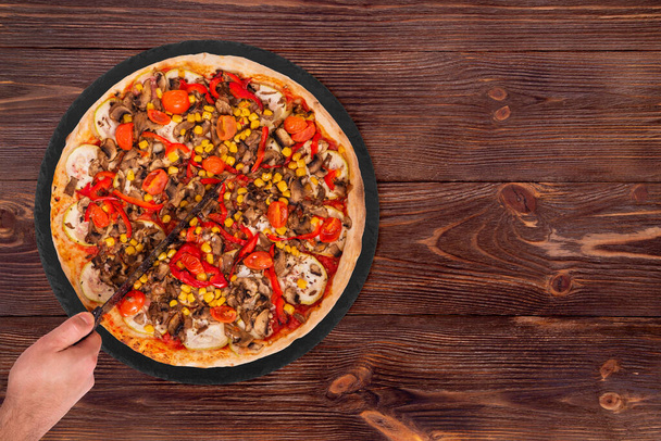 Ο άνθρωπος έκοψε την πίτσα με ένα κουζινομάχαιρο. Πίτσα με μανιτάρια, καλαμπόκι, τομάτα κεράσι, κολοκύθια και πιπεριές ή Veggie Χορτοφαγική πίτσα στο πιάτο σχιστόλιθο σε ξύλινο φόντο, top view - Φωτογραφία, εικόνα