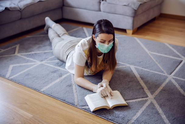 Mujer en aislamiento domiciliario usando mascarilla facial y guantes, leyendo un libro y estudiando información sobre coronavirus outbrake 2020. Educación en el hogar durante la cuarentena
. - Foto, imagen