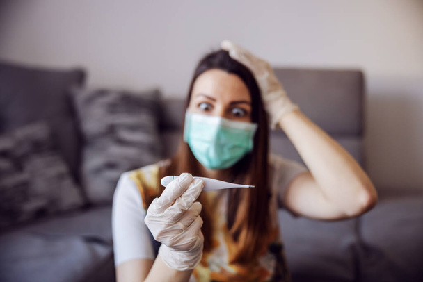 Young woman measuring temperature at home isolation, wearing protective mask and gloves. Stay at home. World pandemic. Symtoms of infection, high temperature, cough and weakness. - Foto, imagen
