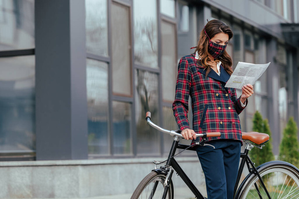 businesswoman in plaid mask standing near bicycle and reading newspaper  - Foto, Imagen