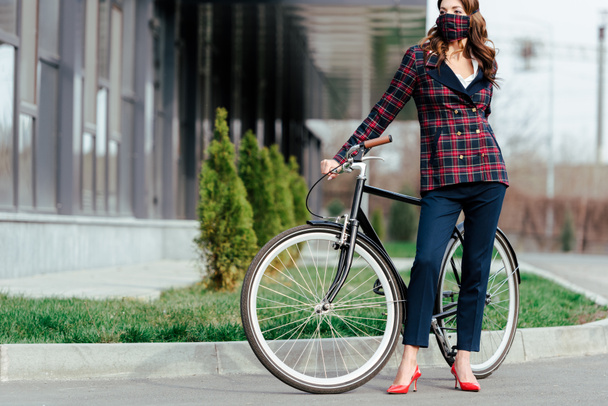 businesswoman in plaid mask standing near bicycle and looking away - Fotó, kép