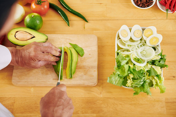 Akşam yemeği için sağlıklı salata yaparken avokado kesen kıdemli adamın elleri. - Fotoğraf, Görsel
