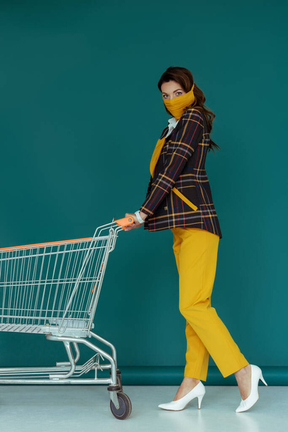 trendy woman in yellow mask walking with empty shopping cart on blue - Foto, Imagem