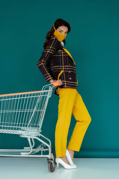 trendy woman in yellow mask posing near empty shopping cart on blue - Fotoğraf, Görsel