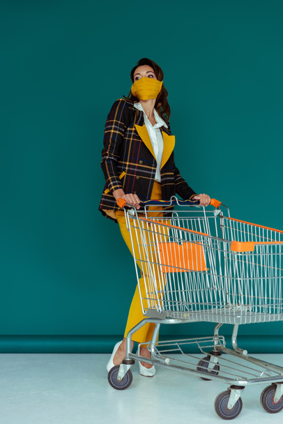 trendy woman in yellow mask standing near empty shopping cart on blue - Fotografie, Obrázek