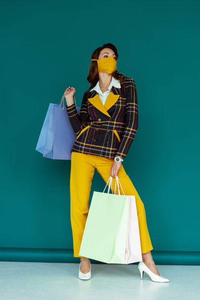 stylish woman in yellow mask and plaid blazer posing with shopping bags on blue - Zdjęcie, obraz