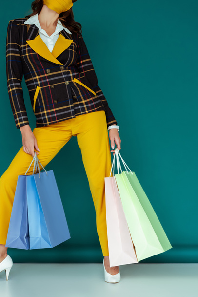 cropped view of stylish woman in yellow mask and plaid blazer posing with shopping bags on blue - Foto, immagini