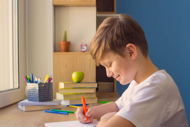 Ευτυχισμένο παιδί αγόρι που γράφει στο βιβλίο του, κάνει εργασίες, σπουδάζει online στο σπίτι στο ξύλινο τραπέζι κοντά στο παράθυρο. Έννοια της ηλεκτρονικής μάθησης - Φωτογραφία, εικόνα