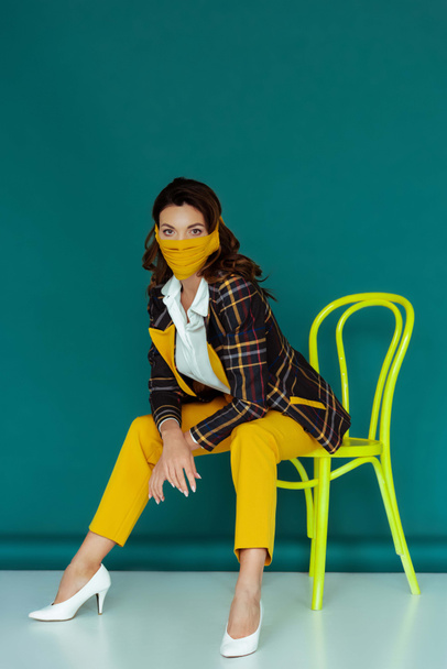stylish woman in yellow mask and plaid blazer posing while sitting on chair on blue - Fotoğraf, Görsel