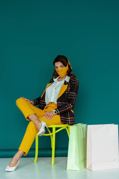 stylish woman in yellow mask and plaid blazer posing while sitting on chair near shopping bags on blue - Fotó, kép