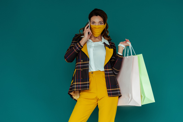 stylish woman in yellow mask and plaid blazer posing with shopping bags and talking on smartphone isolated on blue - 写真・画像