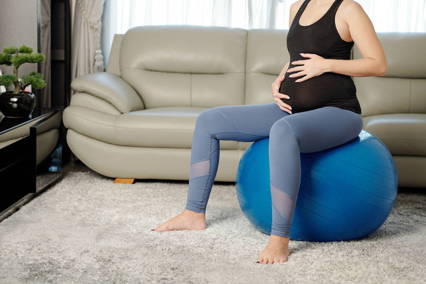 Gewassen beeld van slanke zwangere vrouw aanraken van haar buik wanneer rusten op yoga bal na het sporten - Foto, afbeelding