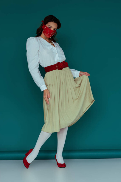 κομψή νεαρή γυναίκα με κόκκινη μάσκα που αγγίζει φούστα σε μπλε  - Φωτογραφία, εικόνα