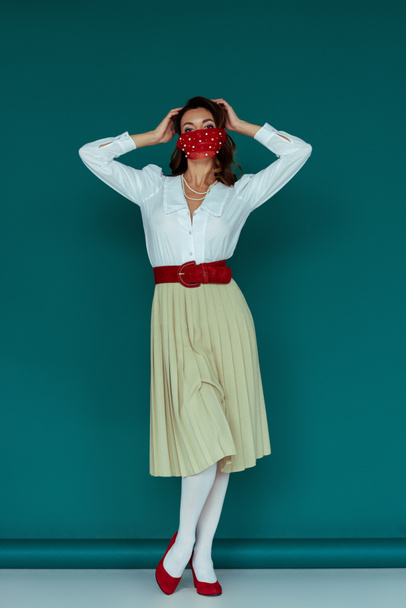stylish young woman in red mask touching hair while posing on blue  - Fotoğraf, Görsel