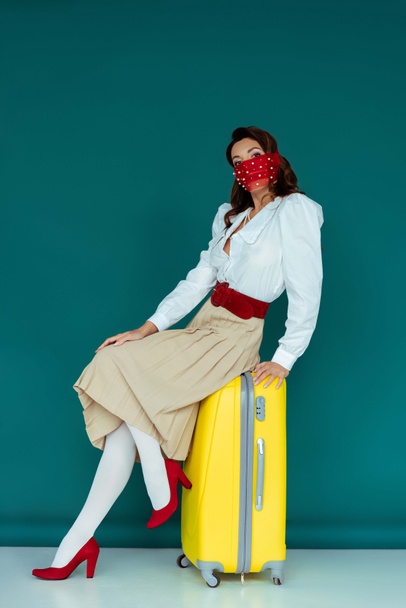 κομψή νεαρή γυναίκα με κόκκινη μάσκα κάθεται σε ταξιδιωτική τσάντα σε μπλε  - Φωτογραφία, εικόνα