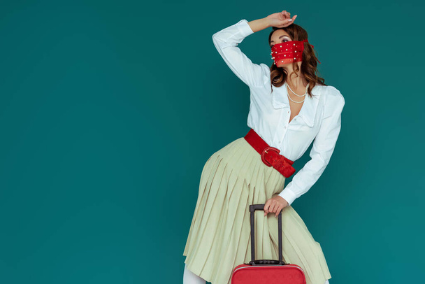 menina elegante em máscara vermelha posando perto de saco de viagem isolado em azul
  - Foto, Imagem