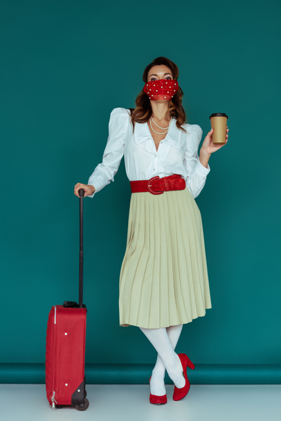 stylish girl in mask holding coffee to go near travel bag on blue  - Foto, afbeelding