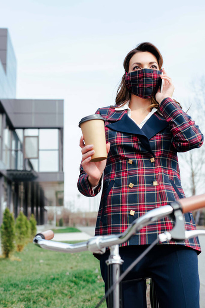 zakenvrouw in ruitmasker houden van koffie te gaan in de buurt van fiets  - Foto, afbeelding