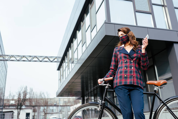 businesswoman in plaid mask standing near bicycle with smartphone  - Foto, Bild