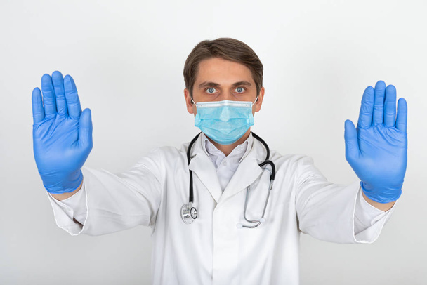 Handsome young physician wearing surgical mask and gloves is showing stop sign on white background - Φωτογραφία, εικόνα