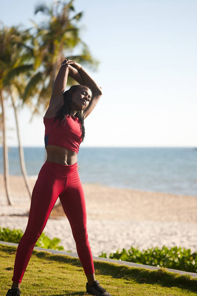 Όμορφη τακτοποιημένη νεαρή μαύρη γυναίκα ζεσταίνεται μετά από τρέξιμο στην παραλία το πρωί - Φωτογραφία, εικόνα