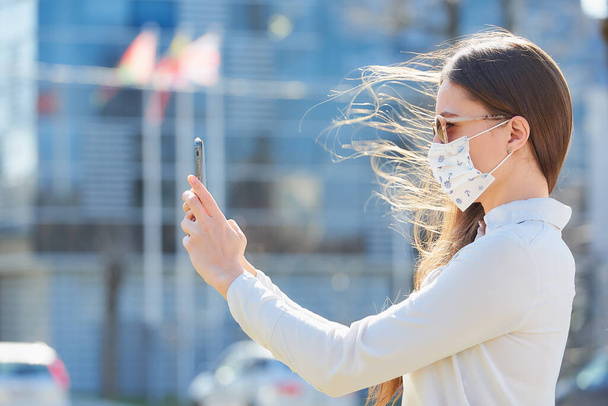 A woman using a smartphone wears a medical face mask to avoid the spread of coronavirus in windy weather. A close-up photo of a lady in sunglasses with a surgical mask on the face against COVID-19. - Valokuva, kuva