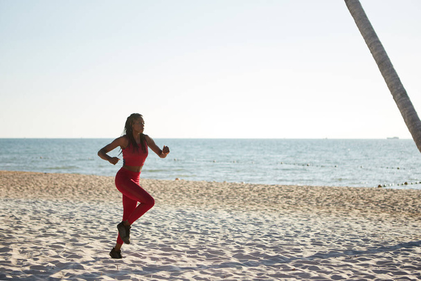 Подходит молодая чернокожая женщина в красном топе и леггинсах, бегущая по песчаному пляжу
 - Фото, изображение