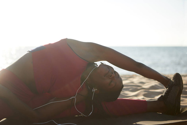 Гибкая спортсменка делает сплиты на песчаном пляже и наклоняется к левой ноге
 - Фото, изображение