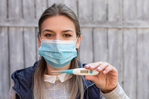 Mujer contaminada con covid-19 está usando máscara quirúrgica y sosteniendo termómetro digital al aire libre
 - Foto, imagen