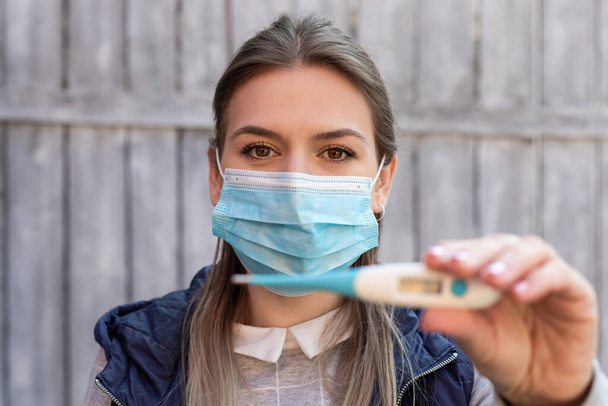 Женщина, зараженная ковид-19, носит хирургическую маску и держит цифровой термометр на открытом воздухе.
 - Фото, изображение