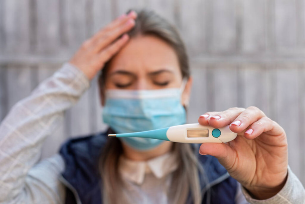 A covid-19-cel fertőzött nő sebészeti maszkot visel és digitális hőmérőt tart a szabadban. - Fotó, kép