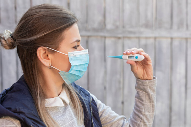 Woman contaminated with covid-19 is wearing surgical mask and holding digital thermometer outdoor - Foto, Imagen