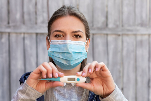 Mujer contaminada con covid-19 está usando máscara quirúrgica y sosteniendo termómetro digital al aire libre
 - Foto, Imagen