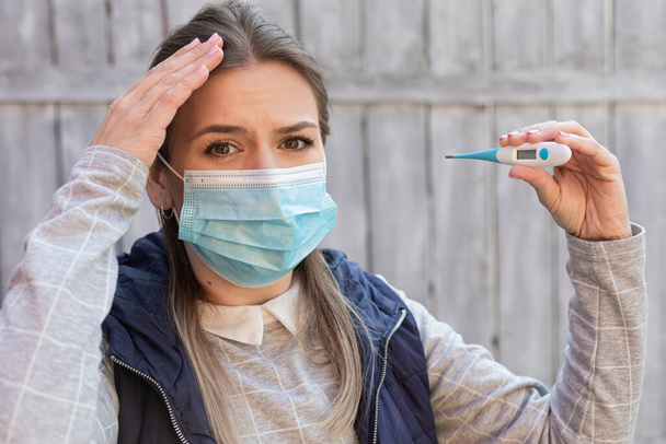 Woman contaminated with covid-19 is wearing surgical mask and holding digital thermometer outdoor - Valokuva, kuva