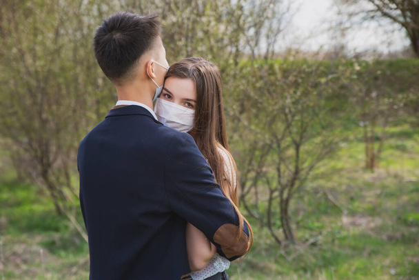 Asian guy with a European girl in medical masks on the background of flowering trees hug and kiss - Foto, Bild