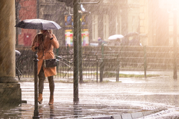 Una mujer con un abrigo marrón bajo un paraguas cruza una calle concurrida bajo fuertes lluvias. Fuertes precipitaciones en la ciudad. Inundación de calles de la ciudad
 - Foto, Imagen